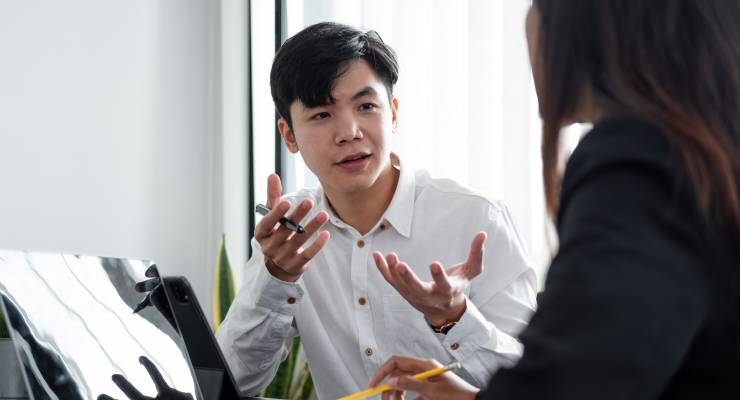 Ein junger Mann ist in einem Coaching-Gespräch mit einer Person. Er trägt ein weißes Hemd und sitzt an einem Schreibtisch mit Laptop vor sich.