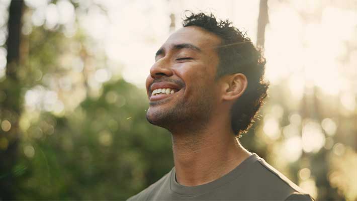 Das Bild zeigt einen lächelnden Mann in der Natur, der entspannt den Kopf zurücklehnt und sinnbildlich für neue Zuversicht und innere Stärke auf dem Weg in eine berufliche Neuorientierung nach Krankheit steht.