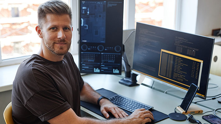 Mann mittleren Alters sitzt an seinem Umschulungsplatz, lächelt in die Kamera und arbeitet an mehreren Bildschirmen mit sichtbarem Programmiercode.
