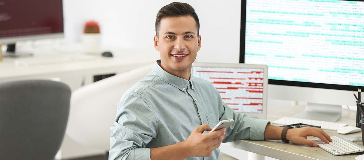Ein Mann sitzt in seiner Online-IT-Administration-Weiterbildung, vor ihm sind ein großer Bildschirm und ein Laptopbildschirm mit Code.
