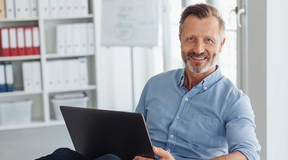 Ein Mann sitzt mit Laptop auf dem Schoß in seinem Büro und findet im Kursnet die passende Weiterbildung.