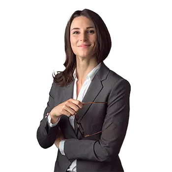 Eine Frau im dunklen Business‑Anzug steht vor hellem Hintergrund und haelt eine Brille in der Hand.