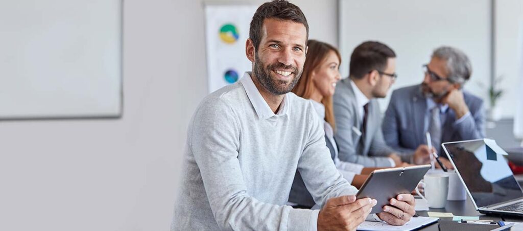 Auf dem Bild ist ein Mann im Vordergrund zu sehen, der ein Tablet in der hand hält. Im Hintergrund sieht man eine Gruppe aus drei Menschen, die sich unterhalten.